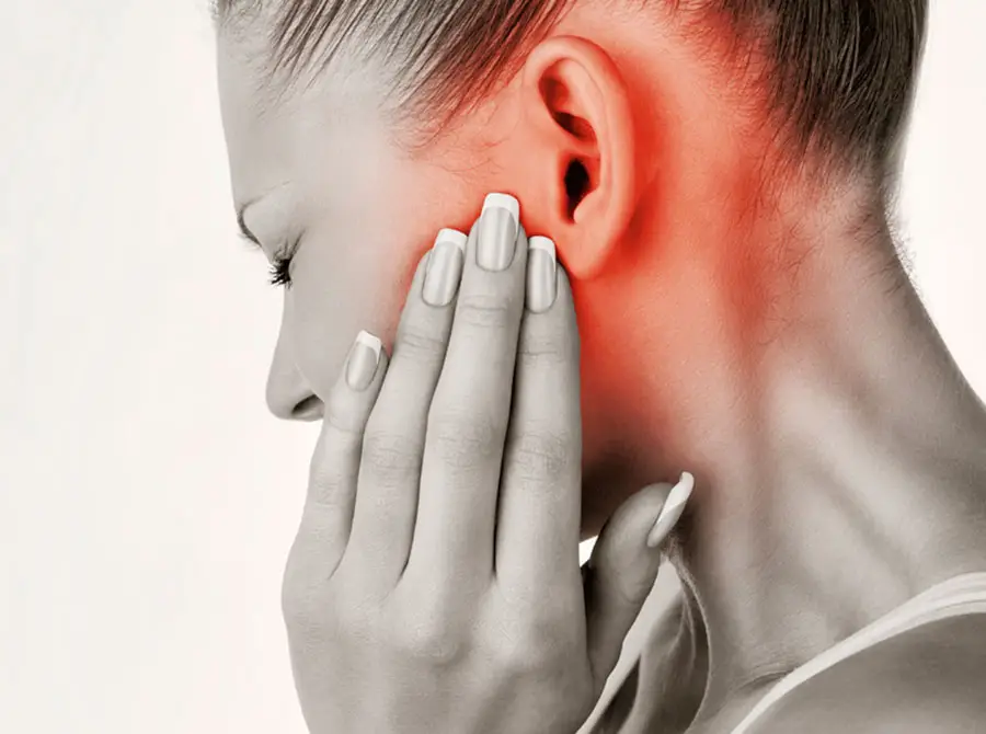 A imagem mostra uma mulher de perfil, tocando suavemente sua bochecha e mandíbula com uma expressão de desconforto. Ela possui cabelos loiros presos e usa unhas pintadas de branco. A área em torno de sua mandíbula é destacada em vermelho para indicar dor, sugerindo que ela pode estar experienciando problemas com sua Articulação Temporomandibular (ATM).